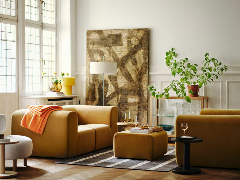 A Stockholm IKEA-inspired living room with mustard yellow sofas, an ottoman, a striped rug, a large abstract painting, plants, and a floor lamp near tall windows.