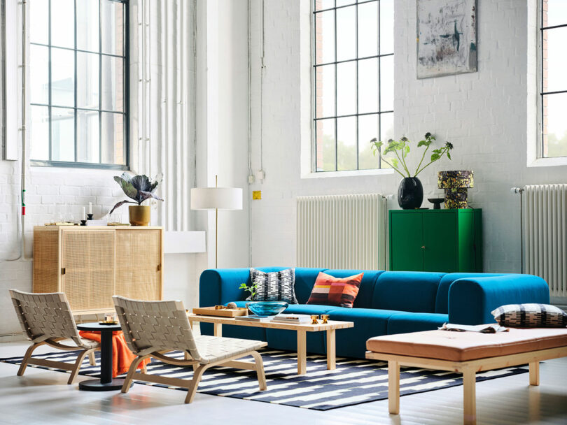 A modern living room with a bright blue Stockholm IKEA sofa, woven chairs, a rectangular coffee table, a green cabinet, indoor plants, and large windows letting in natural light.
