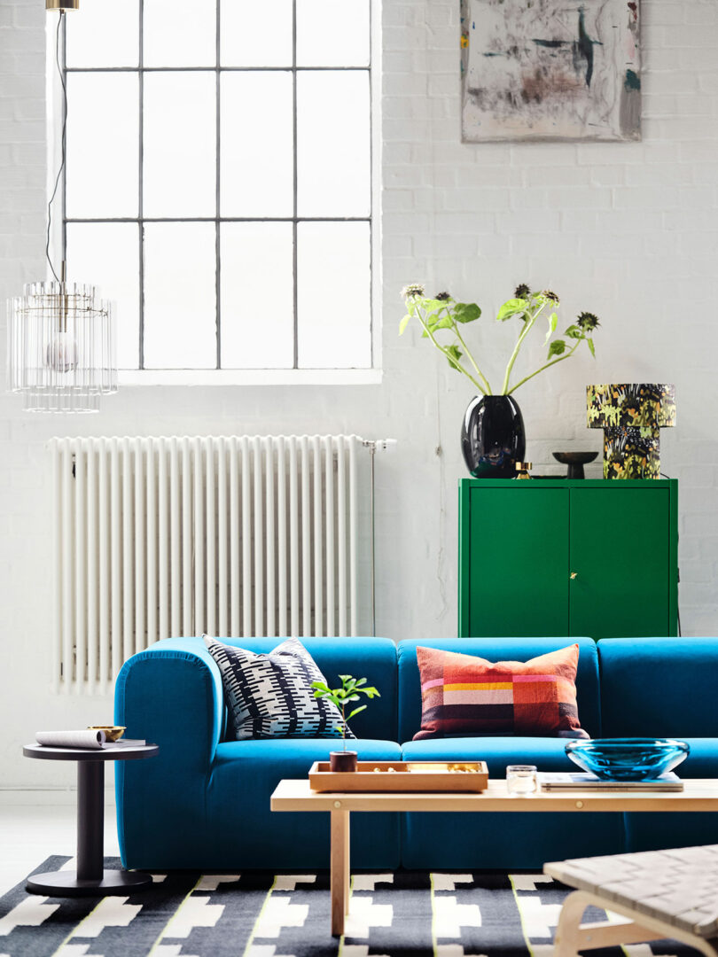 A modern living room inspired by Stockholm IKEA features a blue sofa, colorful cushions, a wooden coffee table, a green cabinet, and a large window letting in natural light.