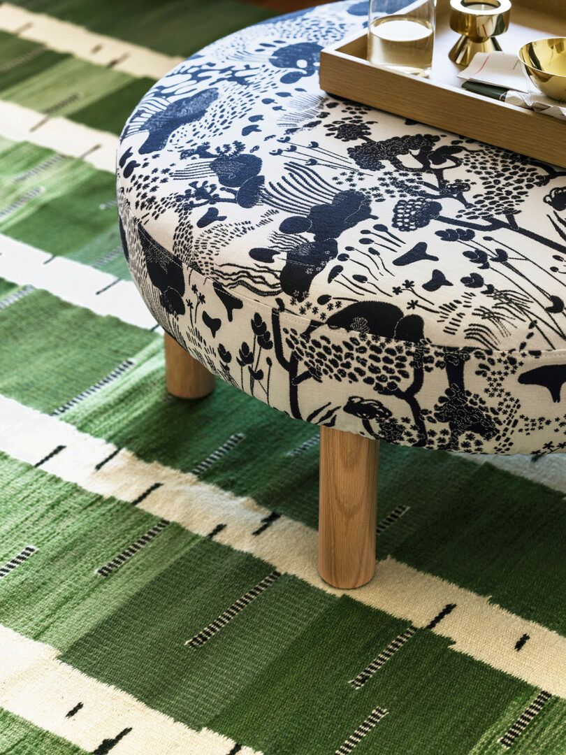 A round, upholstered stool from Stockholm IKEA with a black and white botanical pattern stands on a green patterned rug. A wooden tray with gold cups sits on top of the stool.