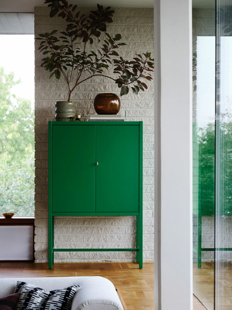 A green Stockholm IKEA cabinet stands against a white brick wall, with decorative vases and a leafy branch placed on top, in a modern, light-filled room.