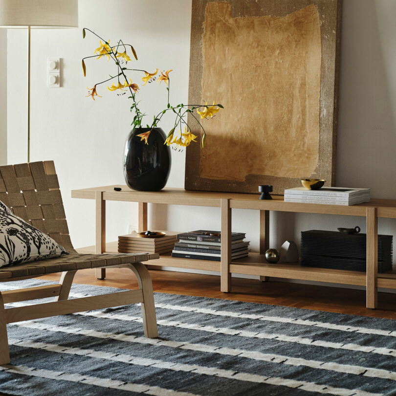 A modern living room with a Stockholm IKEA wooden bench, large abstract artwork, black vase with yellow flowers, woven chair, patterned pillow, and striped rug.