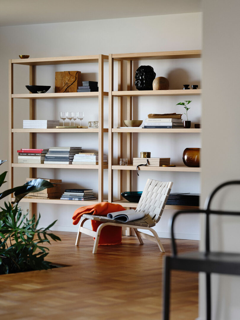 Two wooden bookshelves from Stockholm IKEA, filled with books, ceramics, and glassware, stand behind a white woven lounge chair draped with an orange blanket. The inviting setup sits on a wood floor with a plant in the foreground.