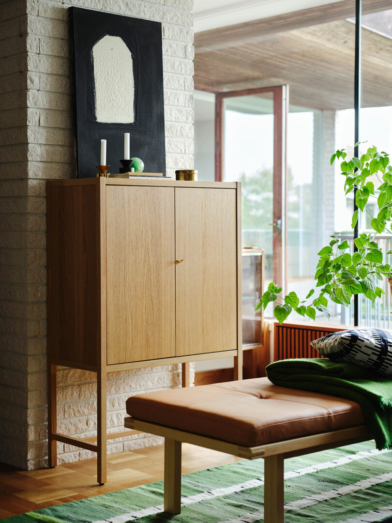 A Stockholm IKEA wooden cabinet stands against a brick wall with candles and decor on top; a framed abstract artwork hangs above. A bench and green plant are in the foreground near large windows.