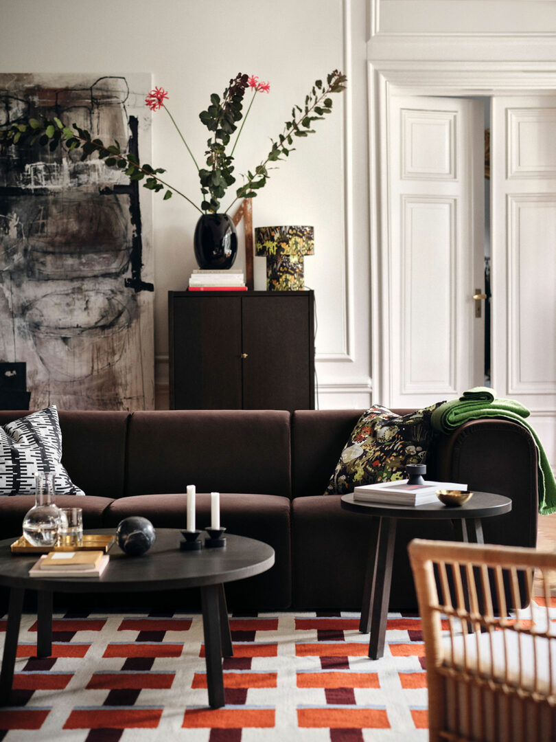 A modern living room with a dark brown sofa, patterned cushions, two round black tables from Stockholm IKEA, candles, decorative books, a vibrant red and cream rug, and a vase with large branches.