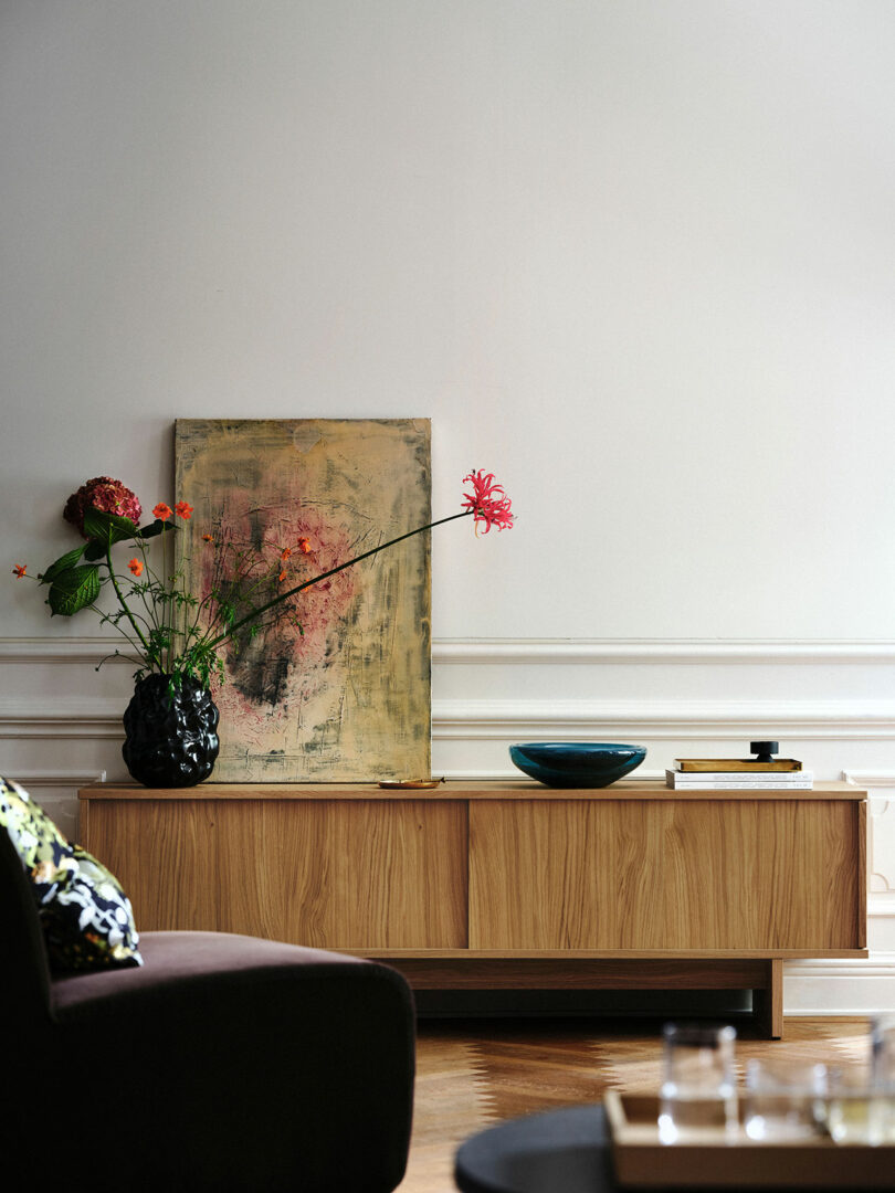 A wooden Stockholm sideboard from IKEA holds a black vase of flowers, a painting, a blue bowl, and a small box against a white wall; part of a dark sofa is visible in the foreground.