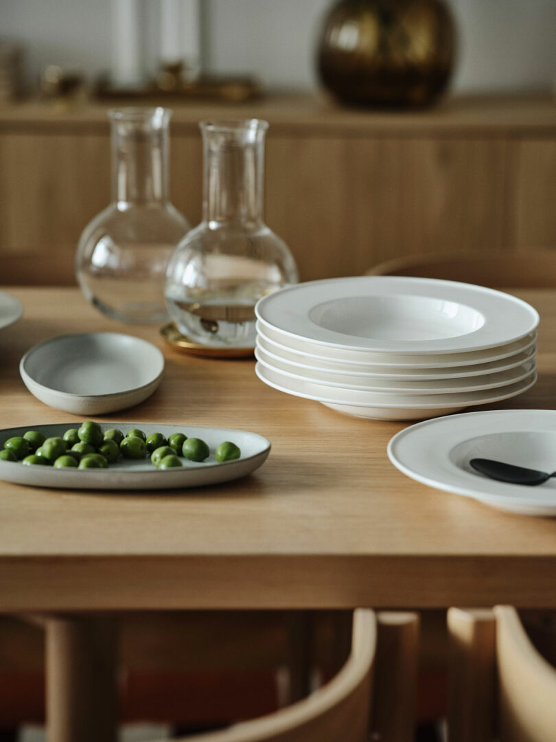A wooden table set with stacked white plates, a dish of green olives, and clear glass carafes brings Stockholm IKEA style to a softly lit dining area.