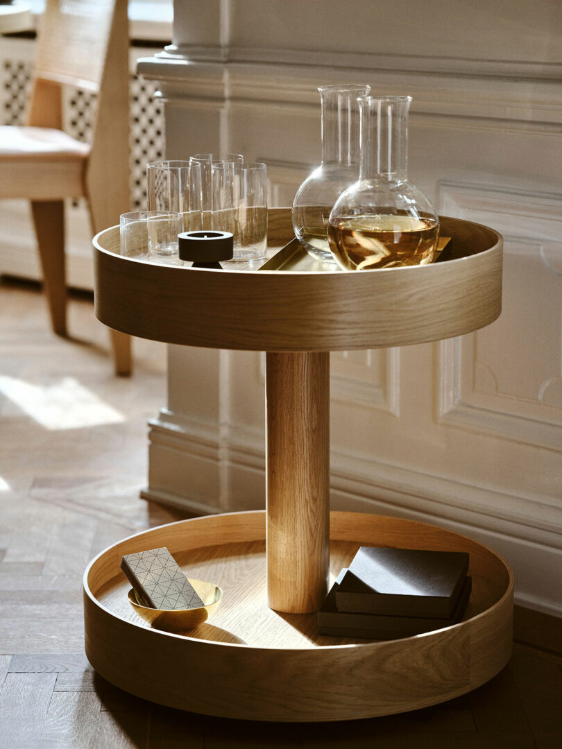 A round wooden Stockholm IKEA bar cart holds glass decanters with amber liquid, drinking glasses, a small bowl, a book, and a chocolate bar, set against a paneled wall.