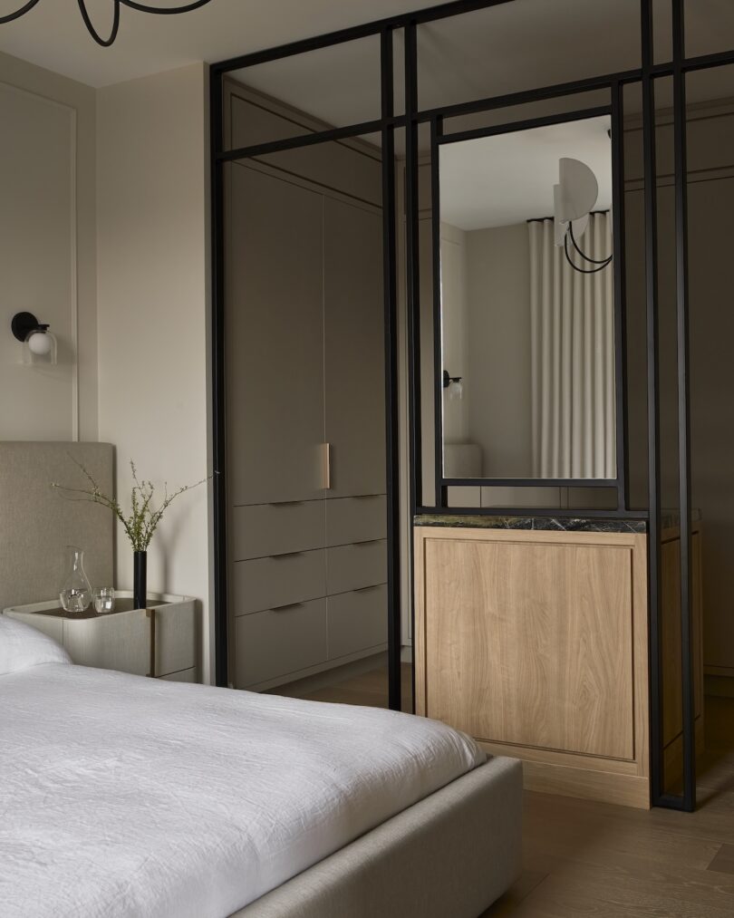 Minimalist bedroom with a bed, light gray bedding, a small side table, and a wooden cabinet. A large black-framed mirror is on the right, and built-in closets are visible in the background.