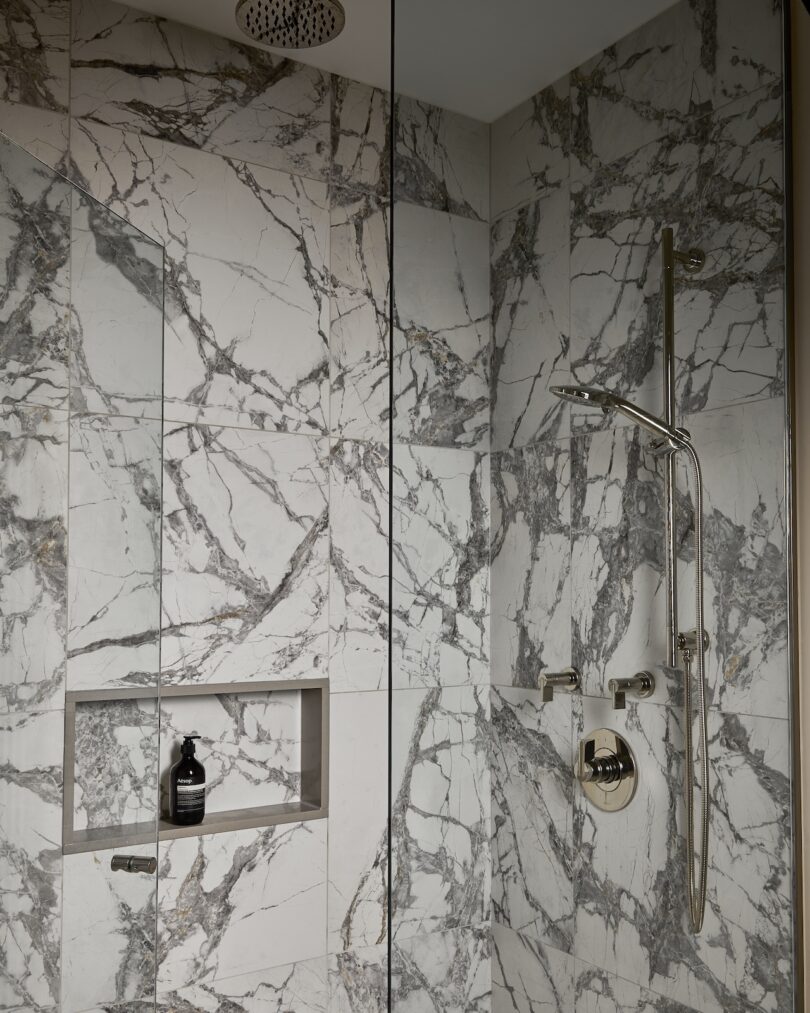 Marble-tiled shower with a rainfall showerhead, a handheld shower fixture, and a built-in niche holding a bottle and soap.
