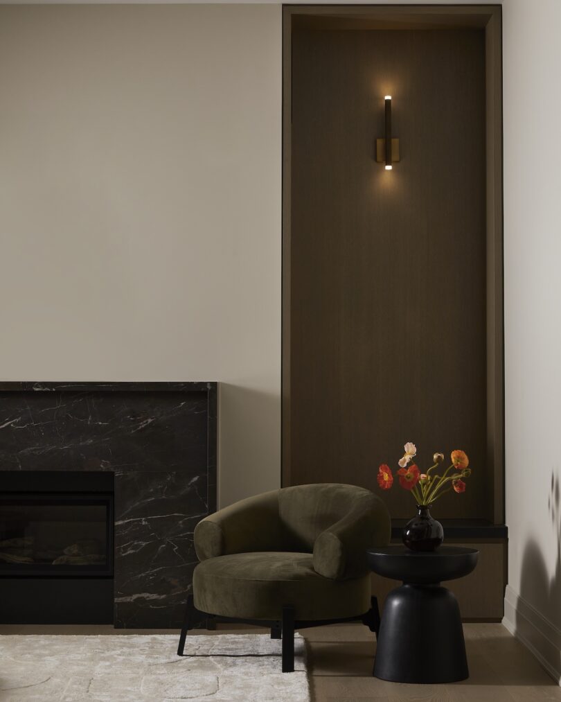 A modern living room corner with a green armchair, small black table with flowers, dark marble fireplace, and a lit wall sconce.