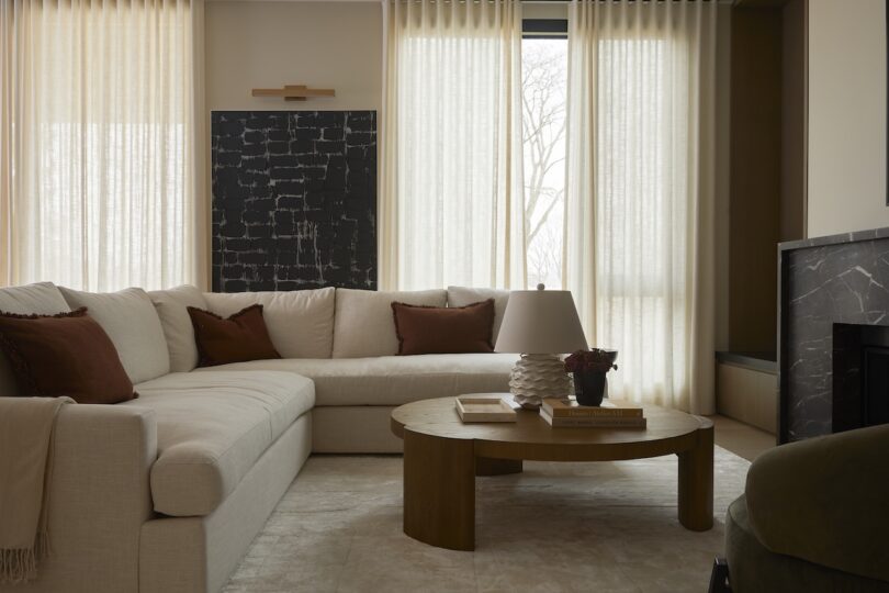 A cozy living room with a large beige sectional sofa, round wooden coffee table, and a fireplace. Neutral curtains and abstract wall art complete the decor. Natural light filters softly through windows.