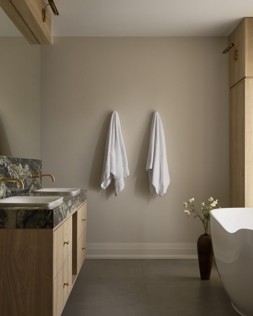Minimalist bathroom with two white towels on the wall, wooden vanity with marble counter, two sinks, gold fixtures, freestanding tub, and a tall vase with flowers.