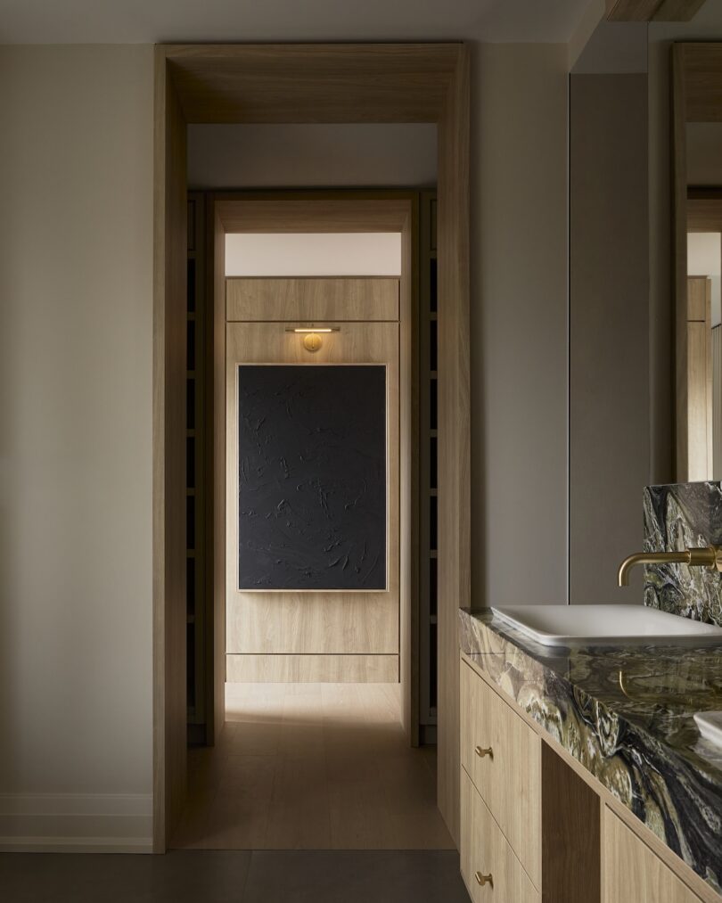 View of a hallway with wood paneling and a black textured artwork on the wall, leading to a bathroom with a marble countertop and a gold faucet.