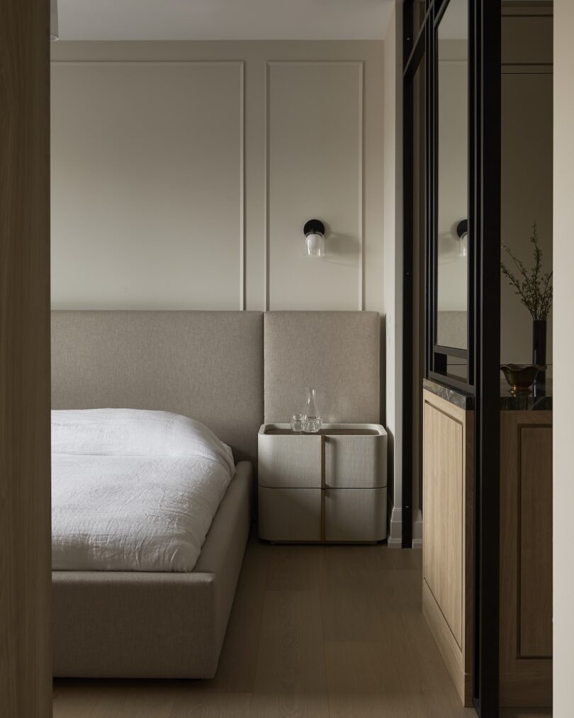 Minimalist bedroom with a beige upholstered bed, a white duvet, and a bedside table holding two glass objects. A wall-mounted black and white light fixture is above the table.
