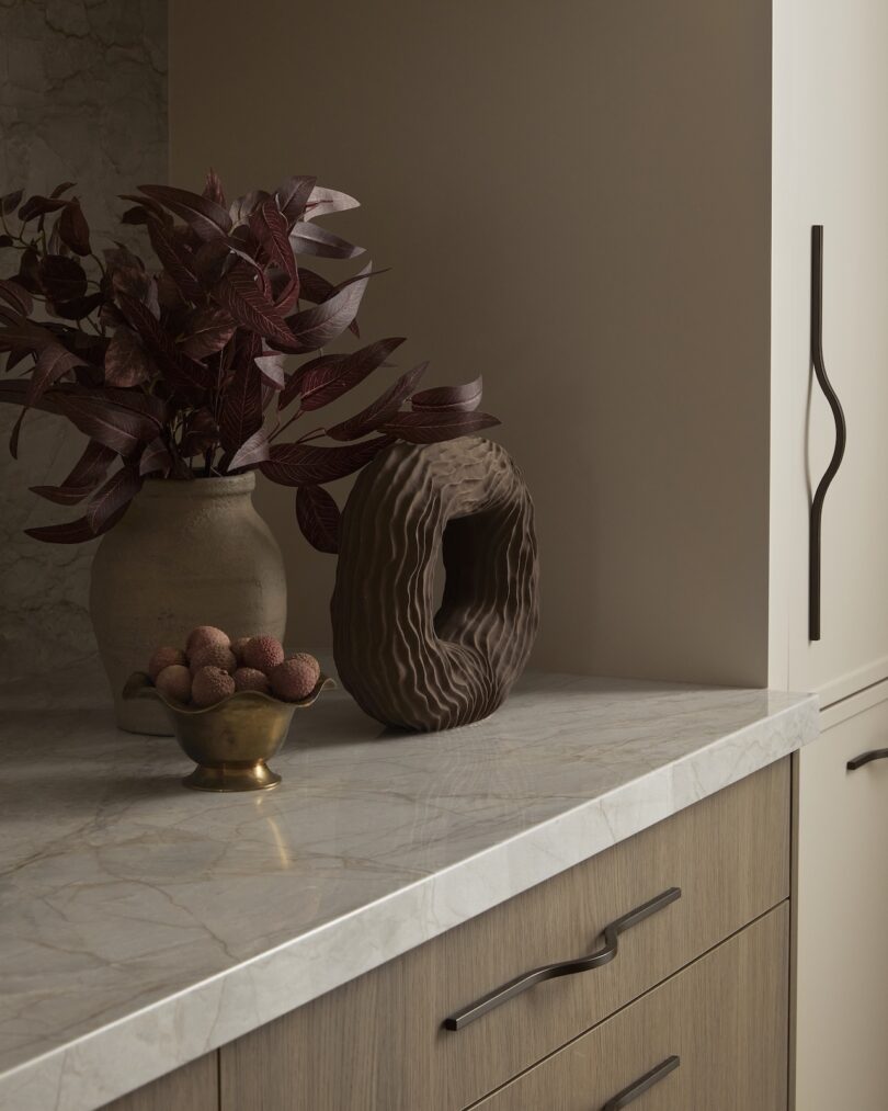 Marble countertop with a decorative brown sculpture, a vase with dark red leaves, and a small brass bowl of round fruits.