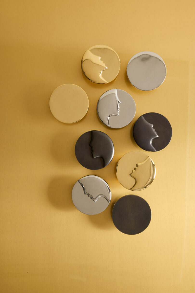 Nine round, metallic containers with face profiles embossed on the lids are arranged on a golden surface. The containers vary in color: gold, silver, and bronze