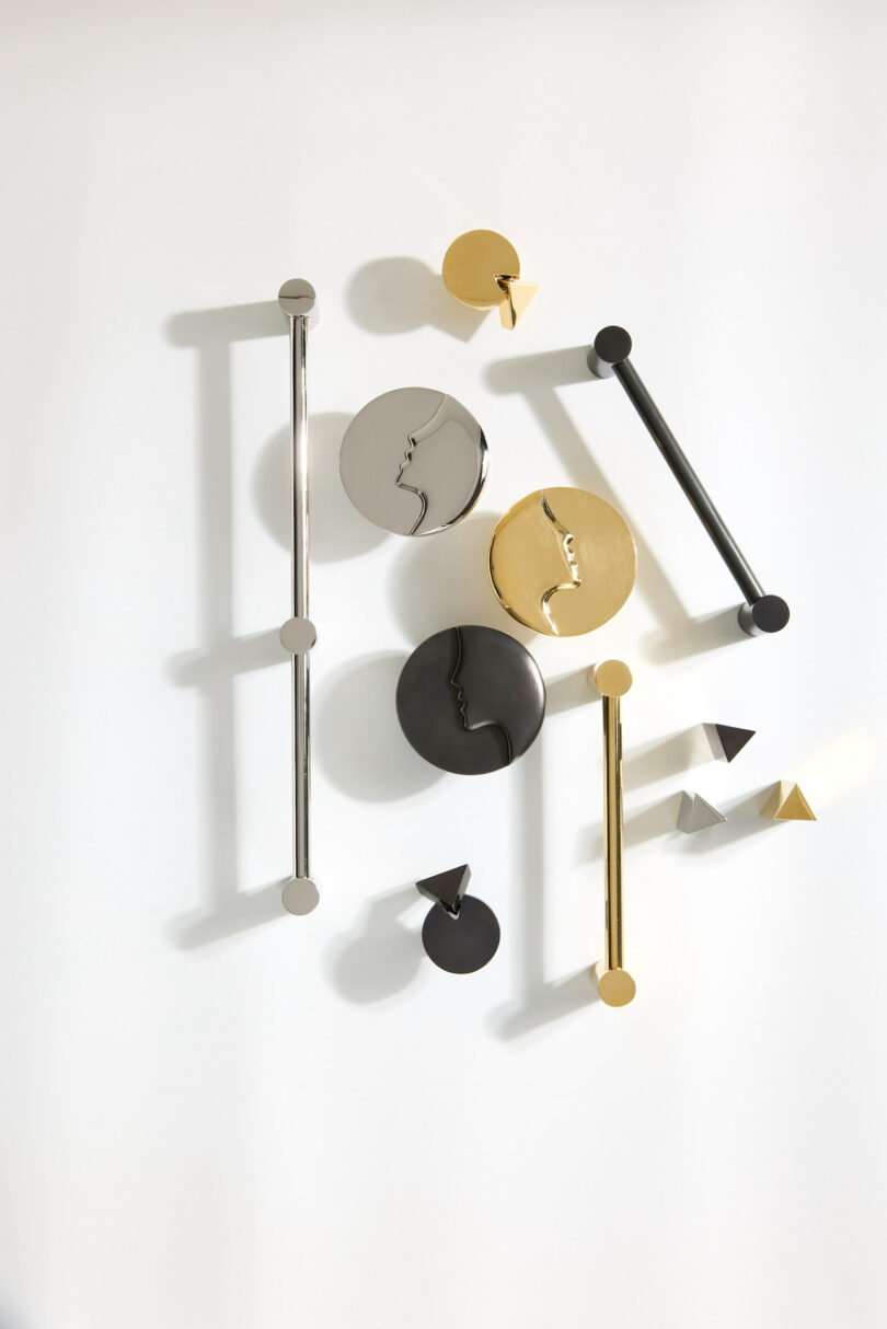 A wall art installation featuring metallic geometric shapes in gold, black, and silver. The composition includes circles, triangles, and lines, casting shadows on a white background.