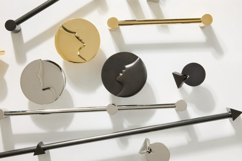 A collection of metal objects shaped like rods and discs with abstract face profiles in gold, silver, and black finishes, arranged on a white surface.
