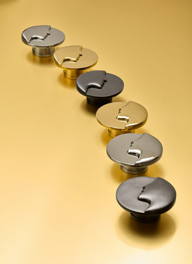 Six round drawer knobs with a face profile design are lined up on a gold surface. They feature different metallic finishes including black, silver, and gold