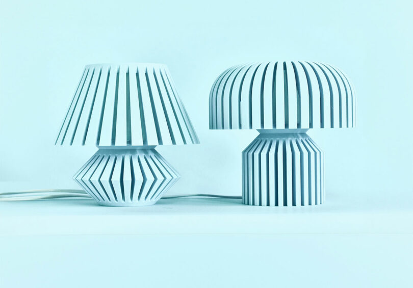 Two modern table lamps from Walmen Dumaliang, with white bases and shades featuring vertical slits, are placed side by side on a white surface against a pale blue background.