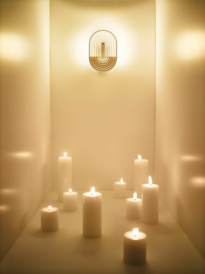 Nine white pillar candles are lit and arranged on a flat surface in a small, beige alcove beneath a modern wall sconce emitting warm light, creating a serene ambiance reminiscent of Walmen Dumaliang’s tranquil design style.