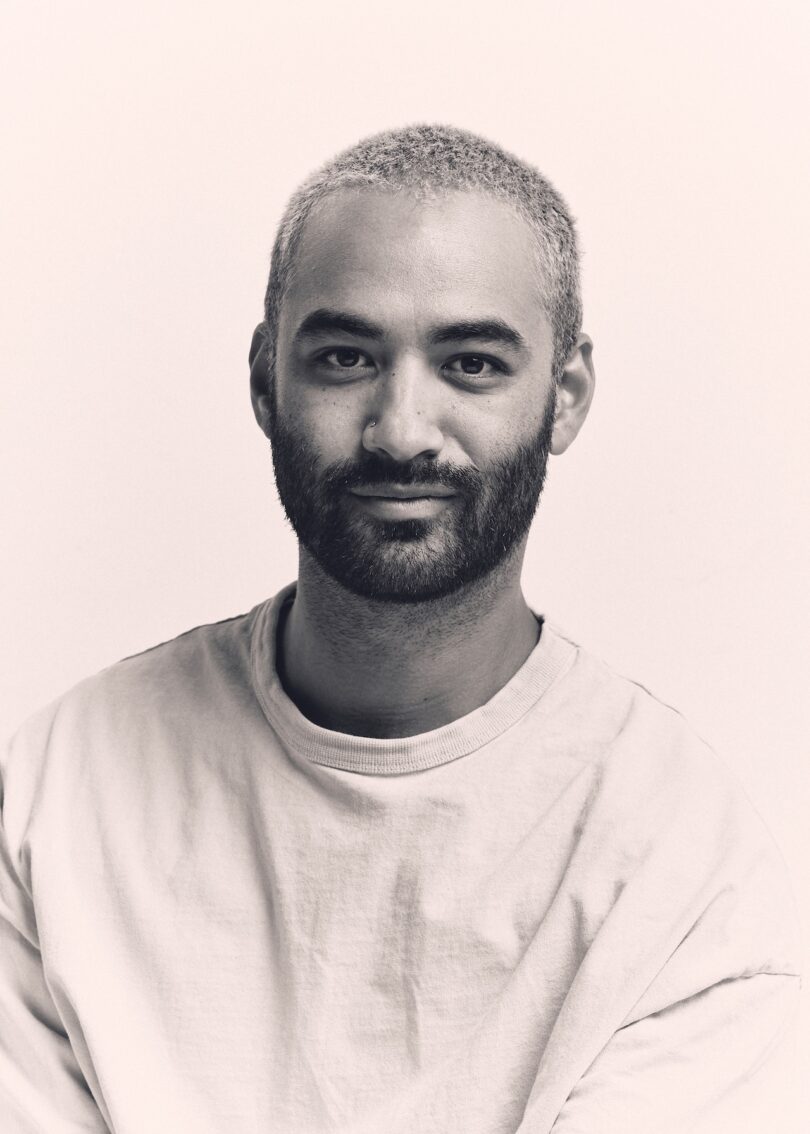A man with short hair and a beard, stands facing the camera in a plain t-shirt, his expression neutral against the light background