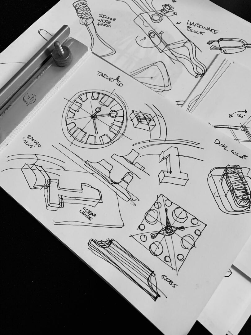 Sketches of various product designs, including a clock face and stylized numbers from the Walmen Dumaliang collection, are drawn in black ink on white paper. Geometric shapes accompany them, with a ruler and pencil nearby for precision