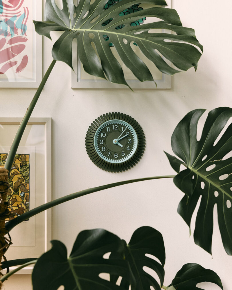 A wall clock with a gear-shaped frame, curated by Walmen Dumaliang, is surrounded by framed art pieces and large Monstera plant leaves