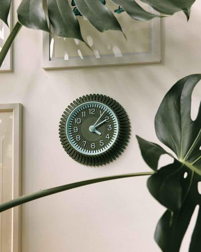 A decorative wall clock with a Walmen Dumaliang sunburst design hangs on a light-colored wall, surrounded by framed art and large green leaves