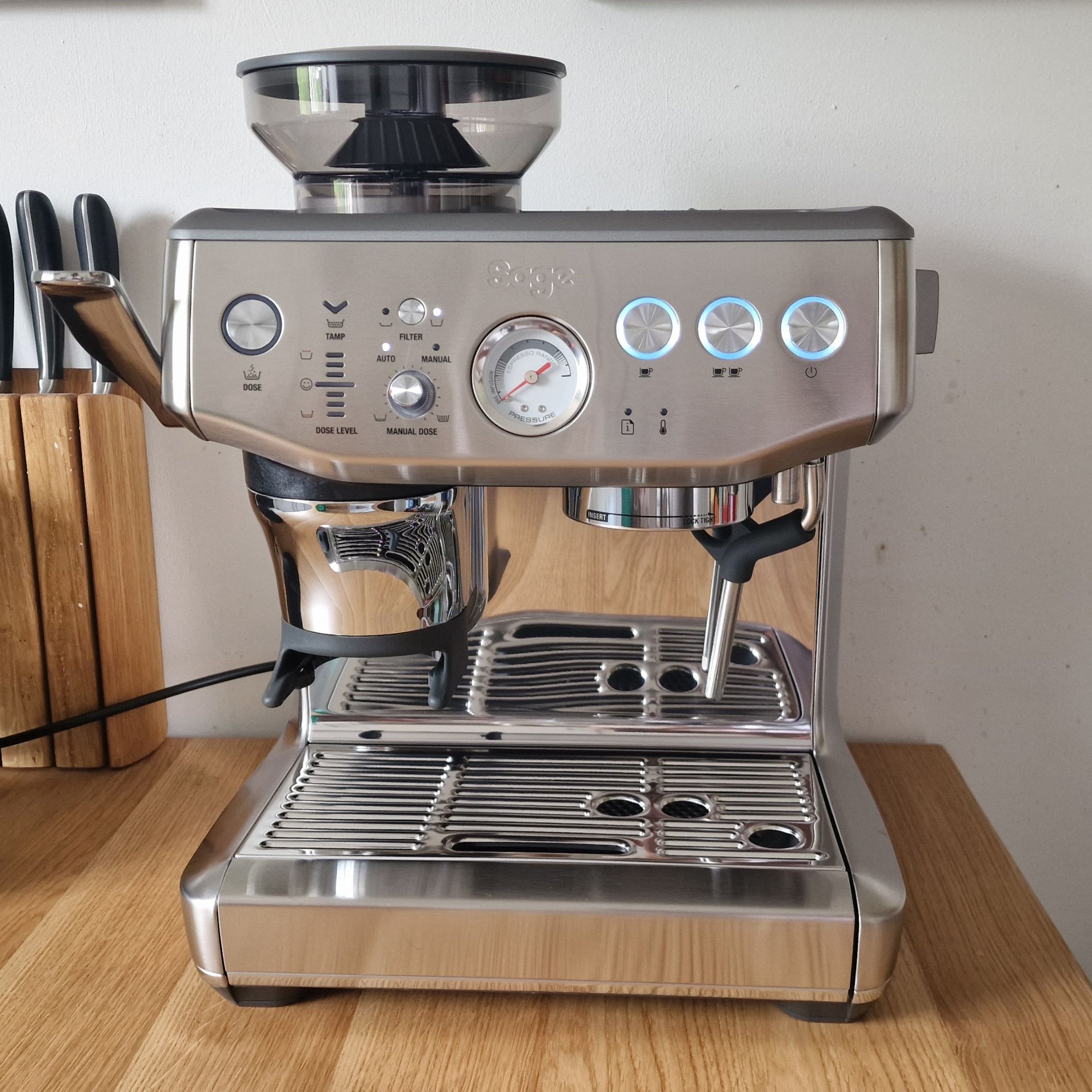 The Sage Barista Express Impress on a wooden surface with a wooden knife block situated behind it