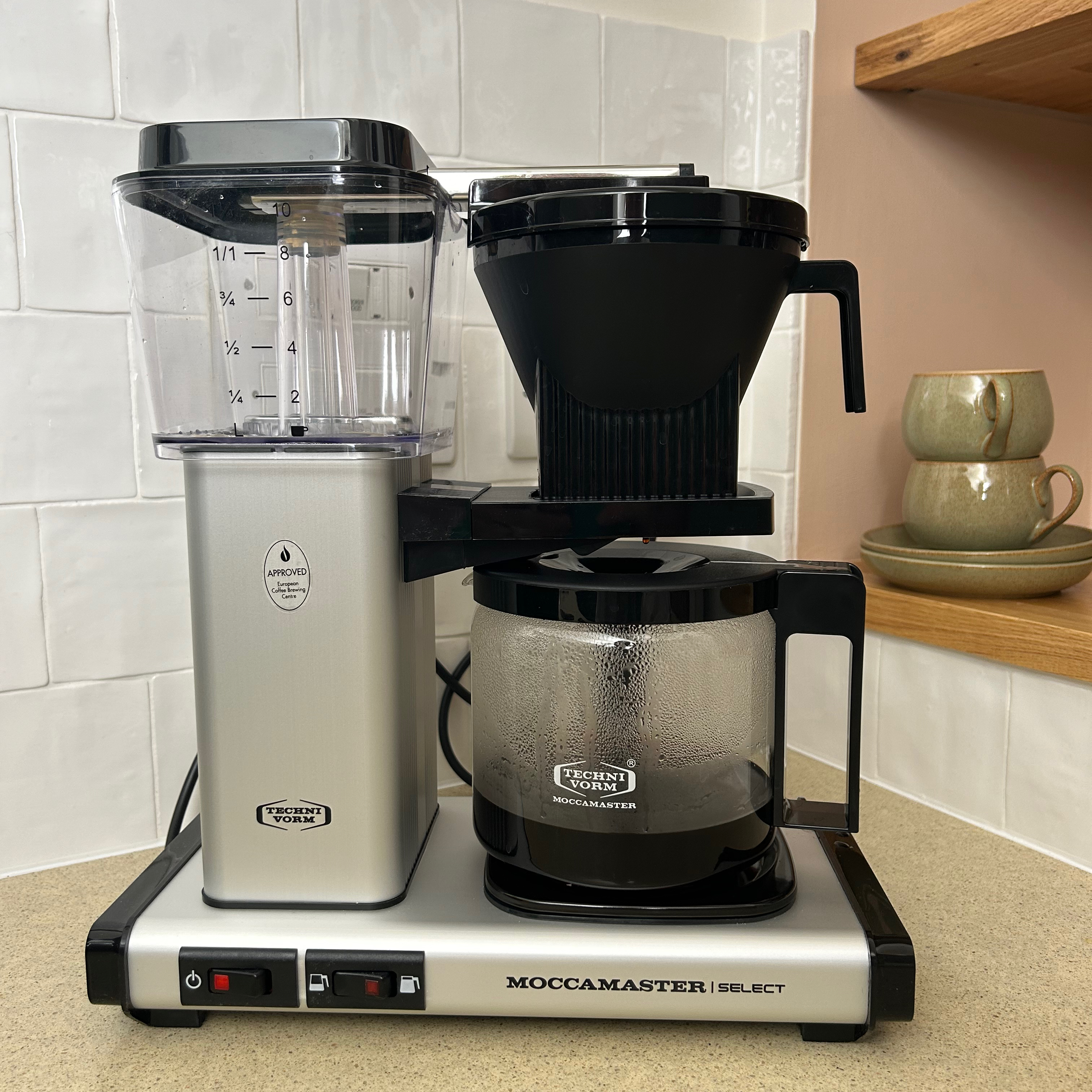 An image of testing the Moccamaster coffee machine at home, situated against a white-tiled wall with two glazed green mugs in the background 