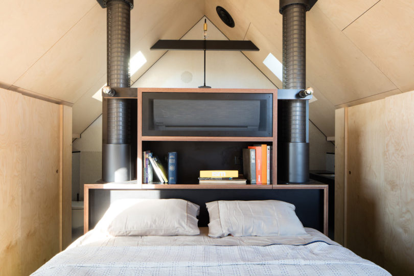 A neatly made bed with two pillows sits against a bookshelf headboard, flanked by black metal pipes in a small, modern lofted room with wood paneling inside a triangular-shaped house.