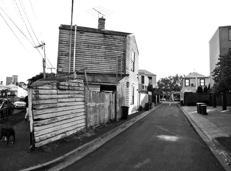 A weathered, triangular-shaped house with a leaning fence stands on the corner of a quiet suburban street; a dog is partially visible on the left.