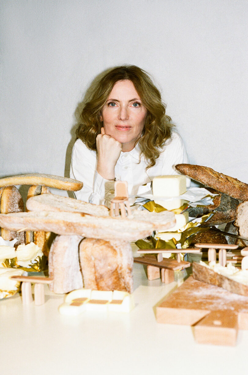 A woman with wavy hair and a white shirt sits at a table displaying breads, cheeses, and other food items arranged in sculptural forms reminiscent of Faye Toogood’s distinctive aesthetic.