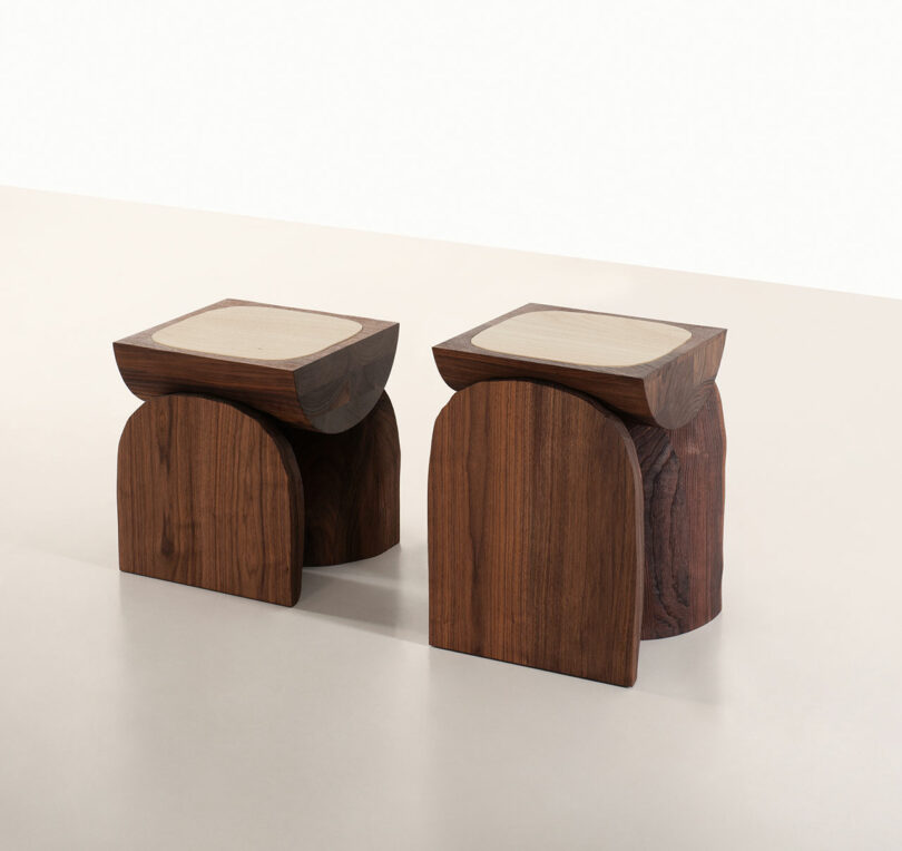 Two wooden stools with curved bases and flat, light-colored seats—designed in the distinctive style of Faye Toogood—are placed side by side on a neutral background.