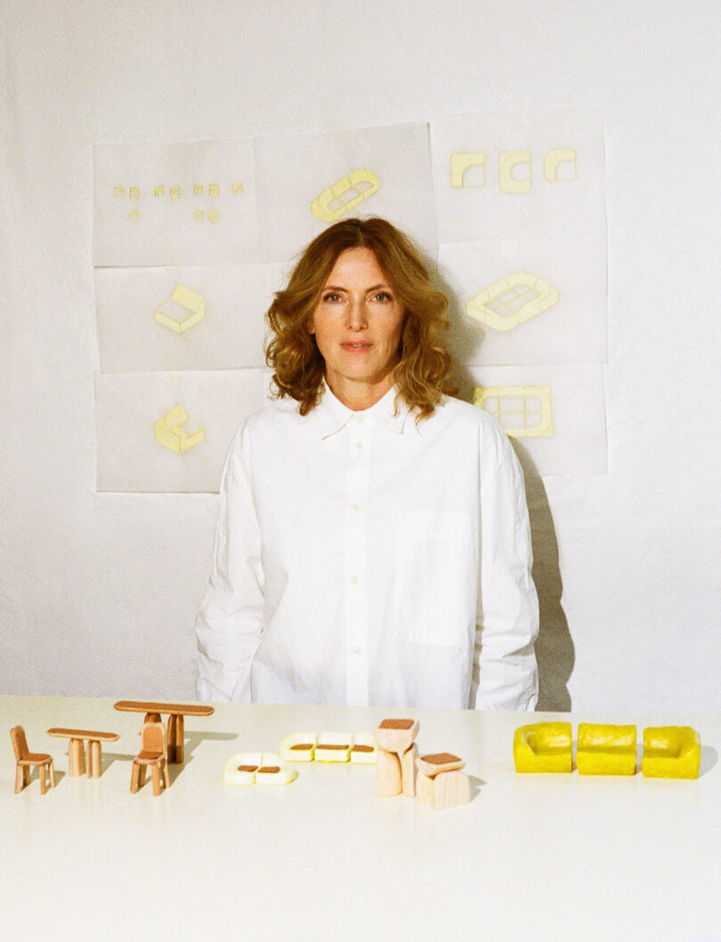 A person in a white shirt stands behind a table displaying small wooden and yellow models, with sketches of Faye Toogood furniture designs on the wall behind them.