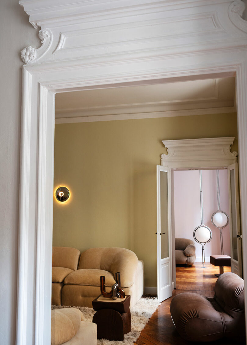 A view through ornate white doorways into a living room with Faye Toogood-inspired beige curved sofas, a round brown table, soft rug, and modern circular wall and floor lights.