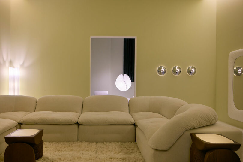 A modern living room with a large beige sectional sofa, shaggy rug, two wooden side tables inspired by Faye Toogood, soft lighting, and round wall sconces on a pale green wall.