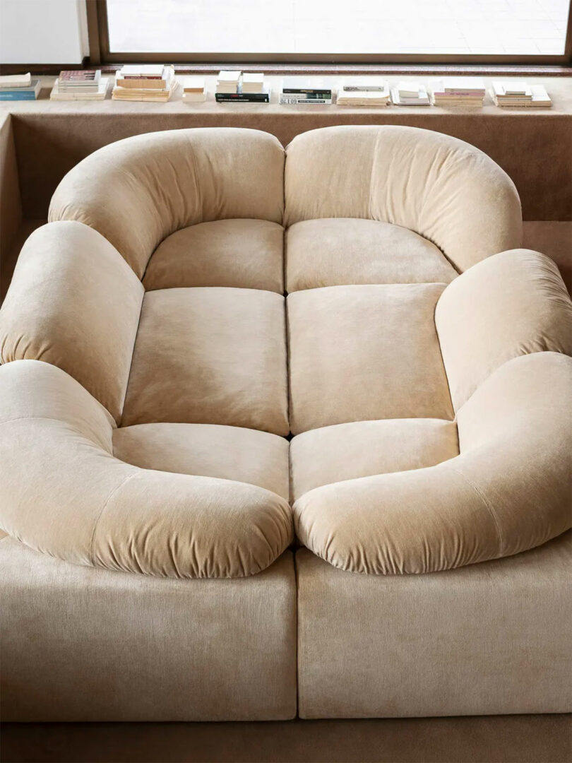 A beige, plush, U-shaped sectional sofa with curved backrests, inspired by Faye Toogood's aesthetic, sits in a modern room with books lined up on a low shelf behind it.