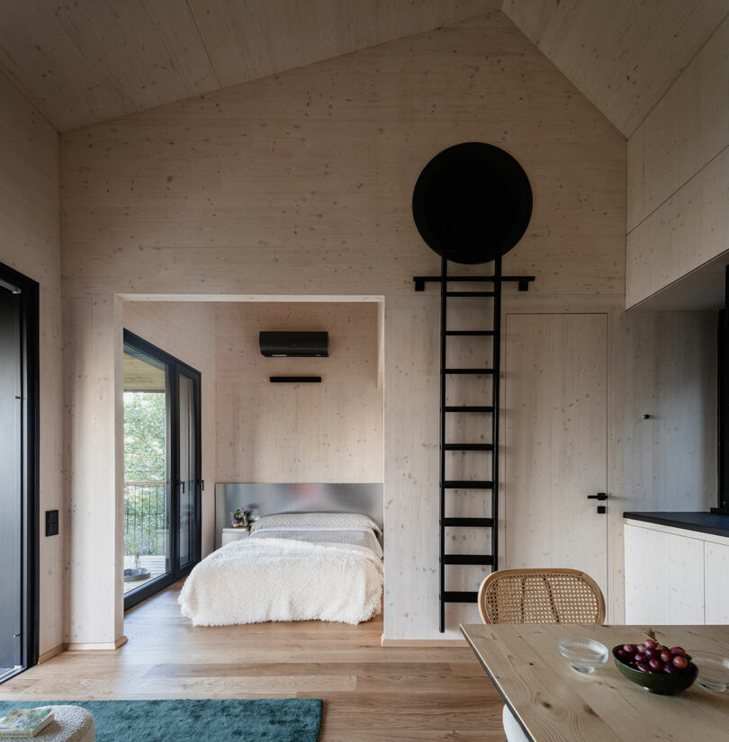 Small modern bedroom with light wood walls, a bed, sliding glass door to a balcony, black metal ladder to a loft, and a dining table with chairs in the foreground.