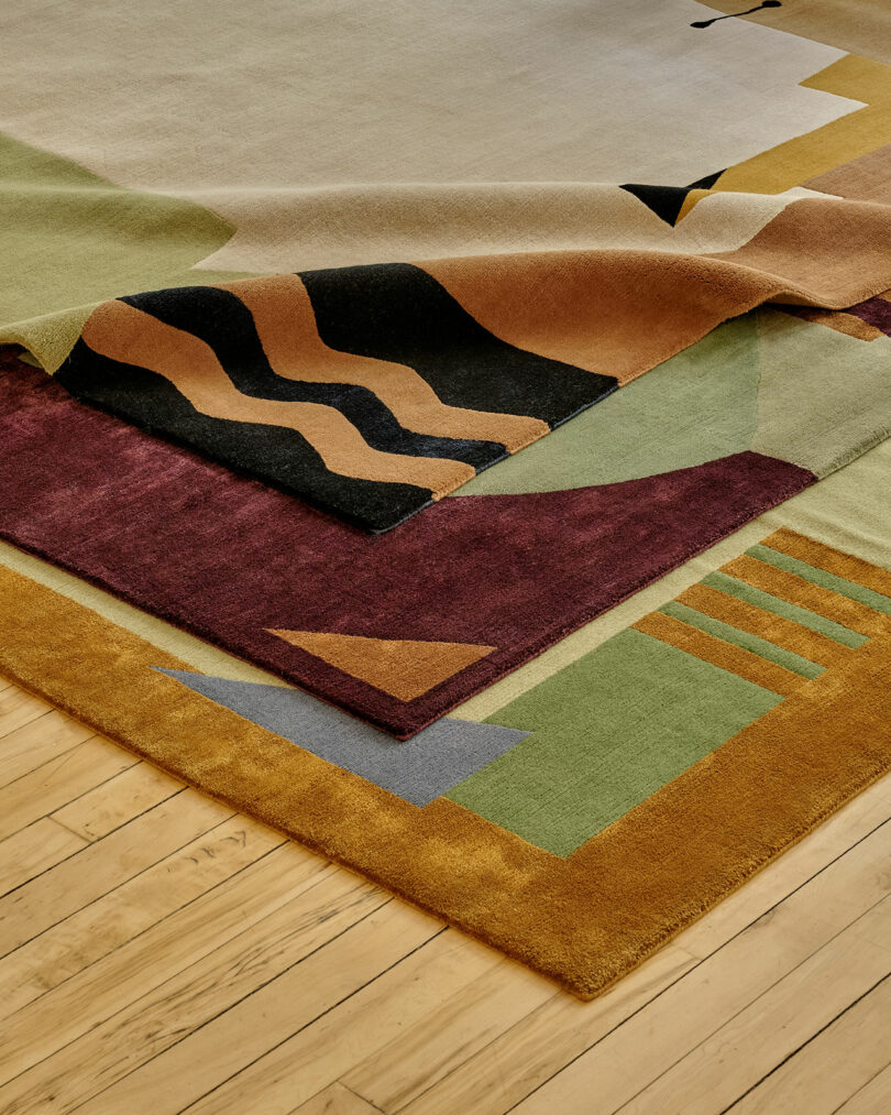 Three colorful, abstract-patterned rugs layered on top of each other on a light wooden floor.
