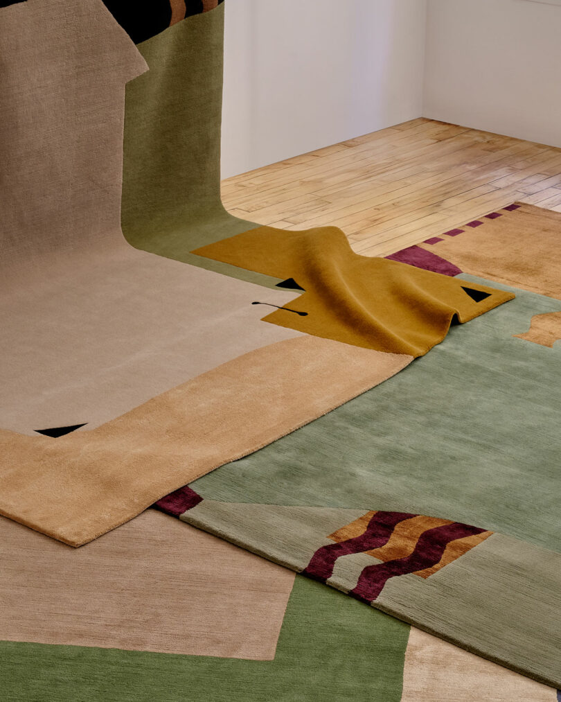 Several abstract-patterned rugs in earthy tones overlap on a light wood floor, with some edges folded and slightly elevated.