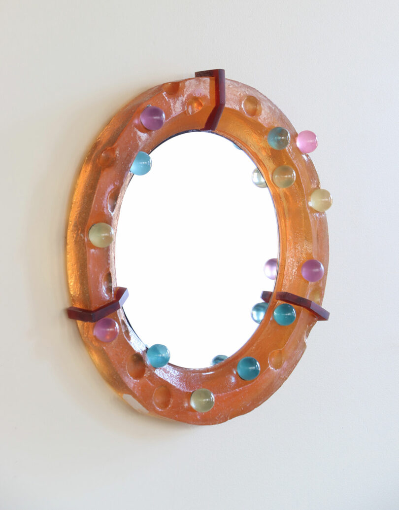 Oval mirror with a translucent orange frame, decorated with evenly spaced colorful round knobs, mounted on a white wall.
