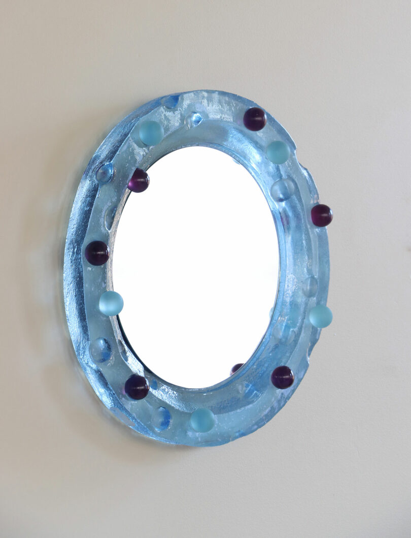 Oval mirror with a thick, translucent blue frame decorated with round blue and purple knobs, mounted on a plain light-colored wall.
