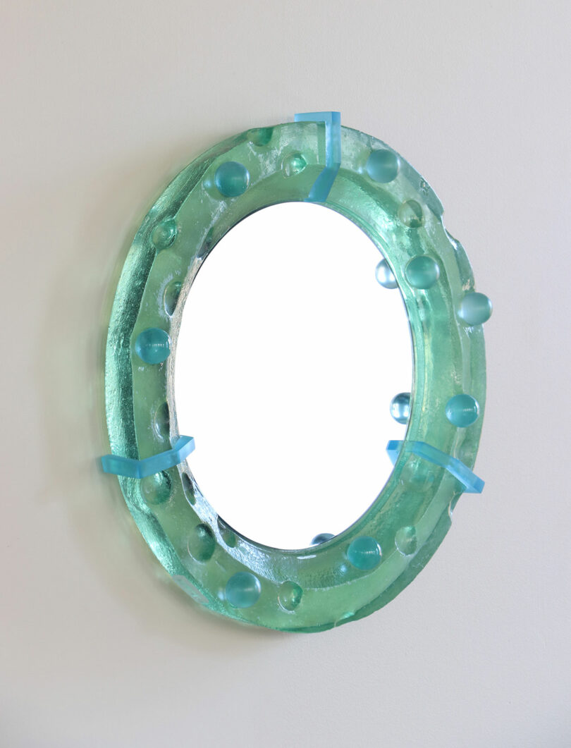 Oval mirror with a thick, translucent green frame featuring raised circular bumps and blue accents, mounted on a white wall.