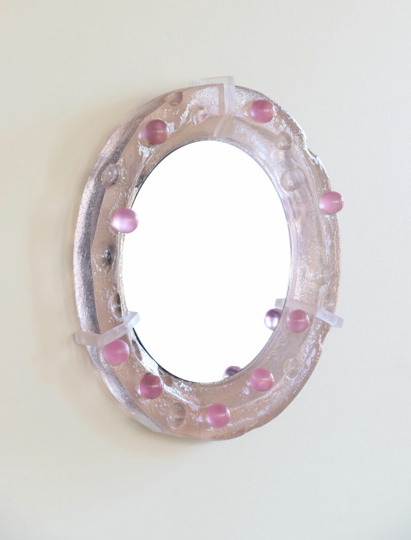 A round mirror with a clear, textured frame decorated with evenly spaced pink spheres is mounted on a plain light-colored wall.