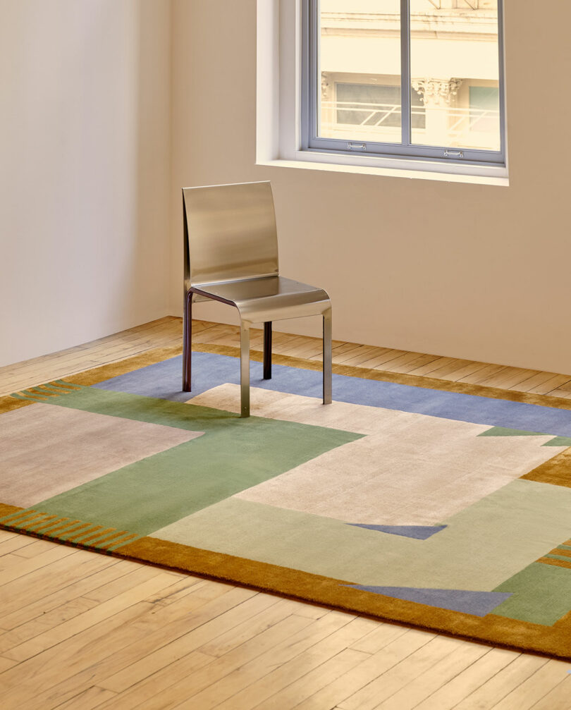 A metal chair sits on a geometric-patterned rug in a room with wooden floors, neutral walls, and a window showing part of a building outside.