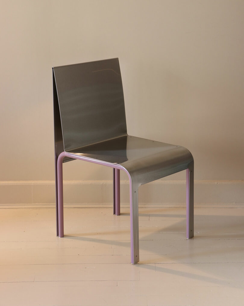 A modern chair with a metallic seat and backrest, featuring curved edges and light purple legs, placed on a light-colored wooden floor against a beige wall.