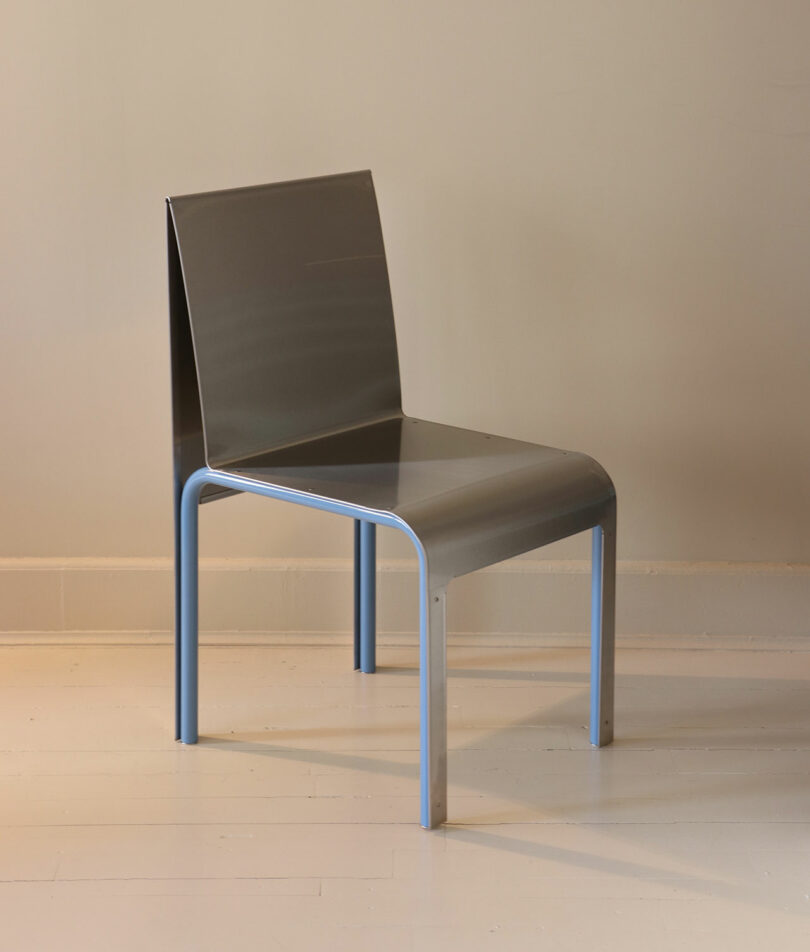 A minimalist metal chair with a reflective seat and backrest, and light blue legs, positioned on a light-colored floor against a beige wall.